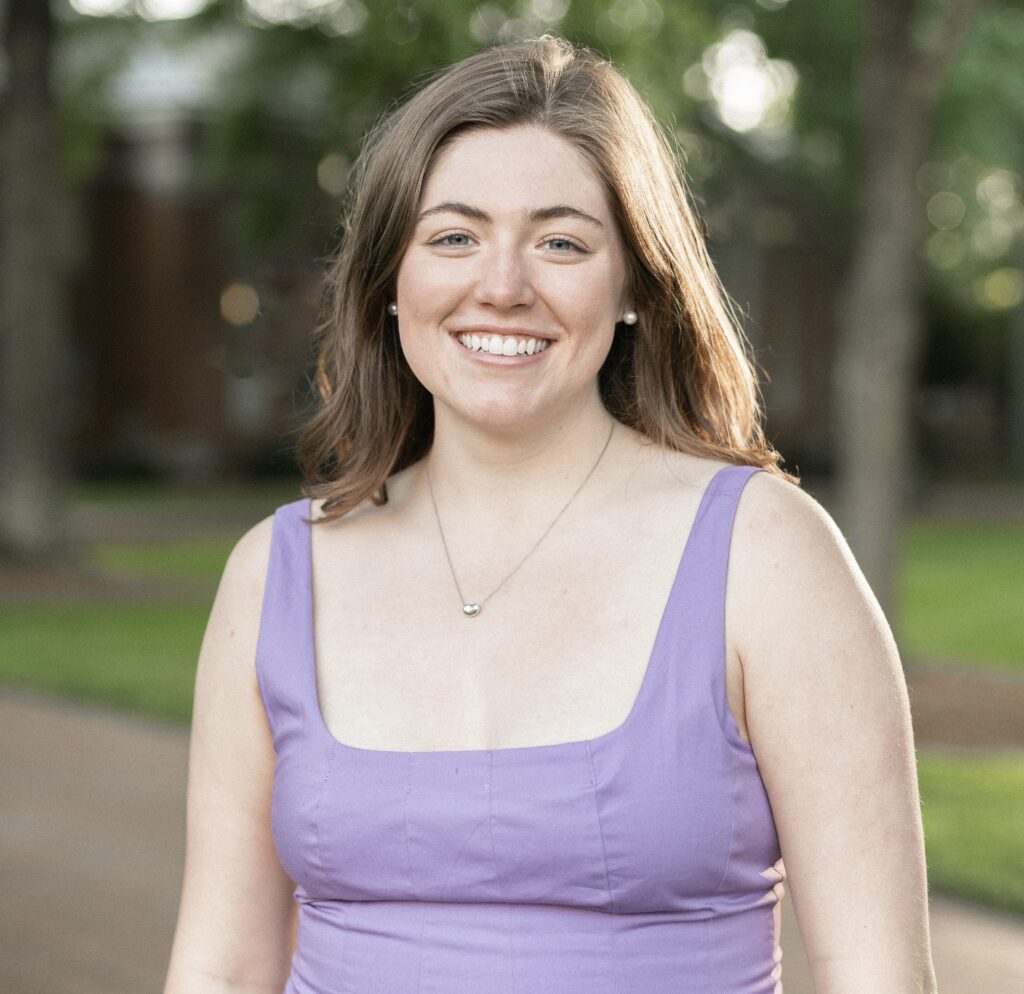 A white woman (Emily Schmitt) with brown hair and a purple dress smiles at the camera.