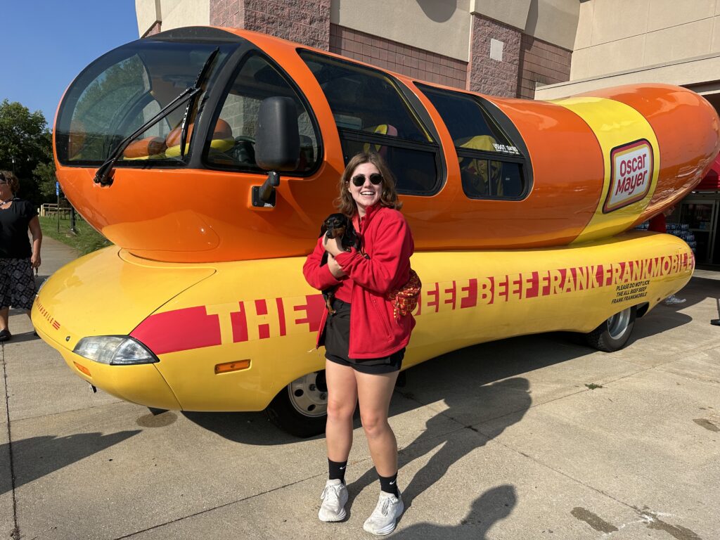 A woman in a red jacket holds and brown and black wiener dog in front of the Wienermobile.