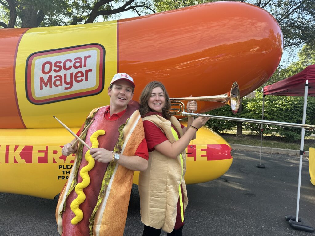 A woman and man hold instruments in front of the wienermobile while wearing hotdog costumes